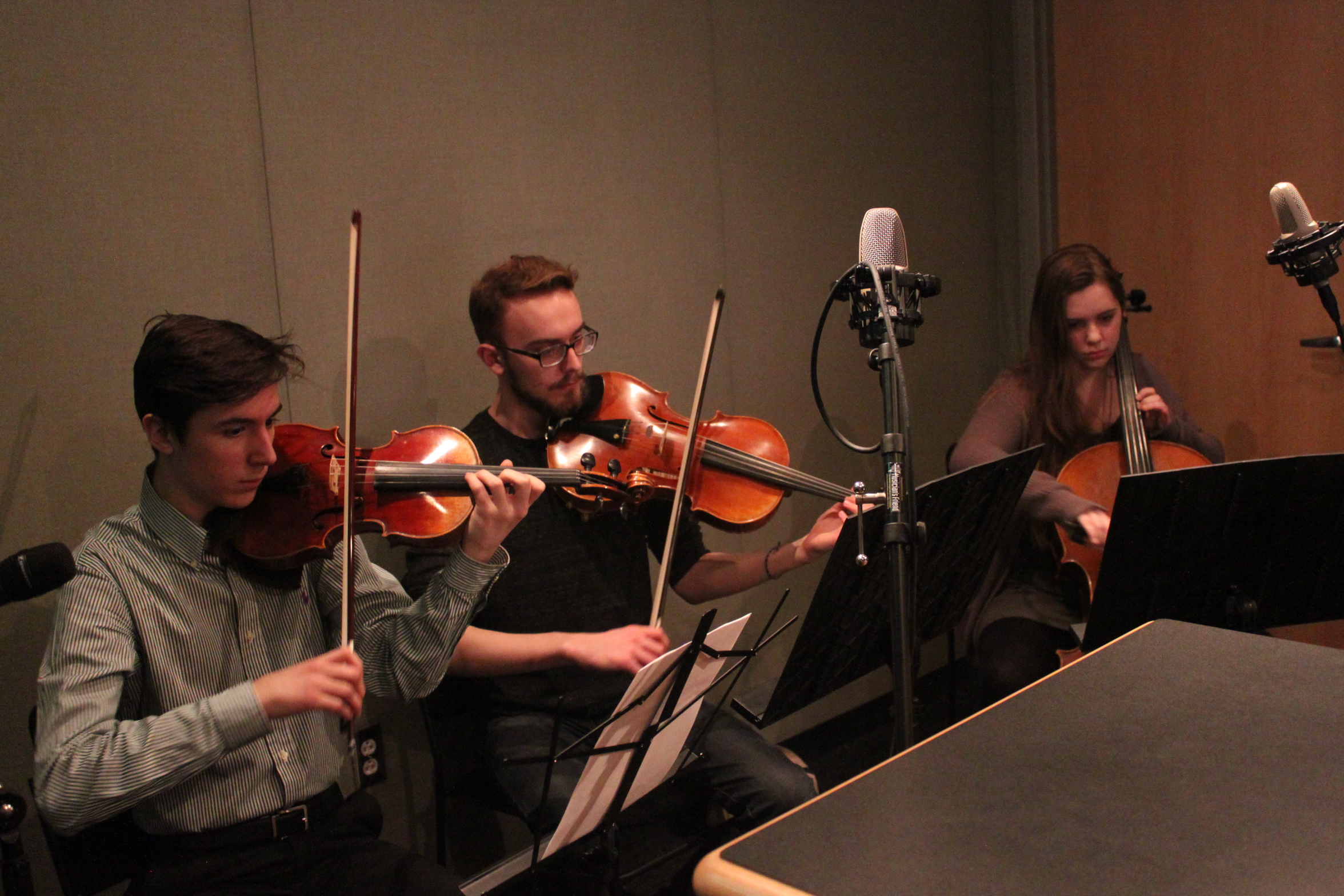 Two teens playing violin and one teen playing cello, all looking at sheet music on music stands in a radio studio