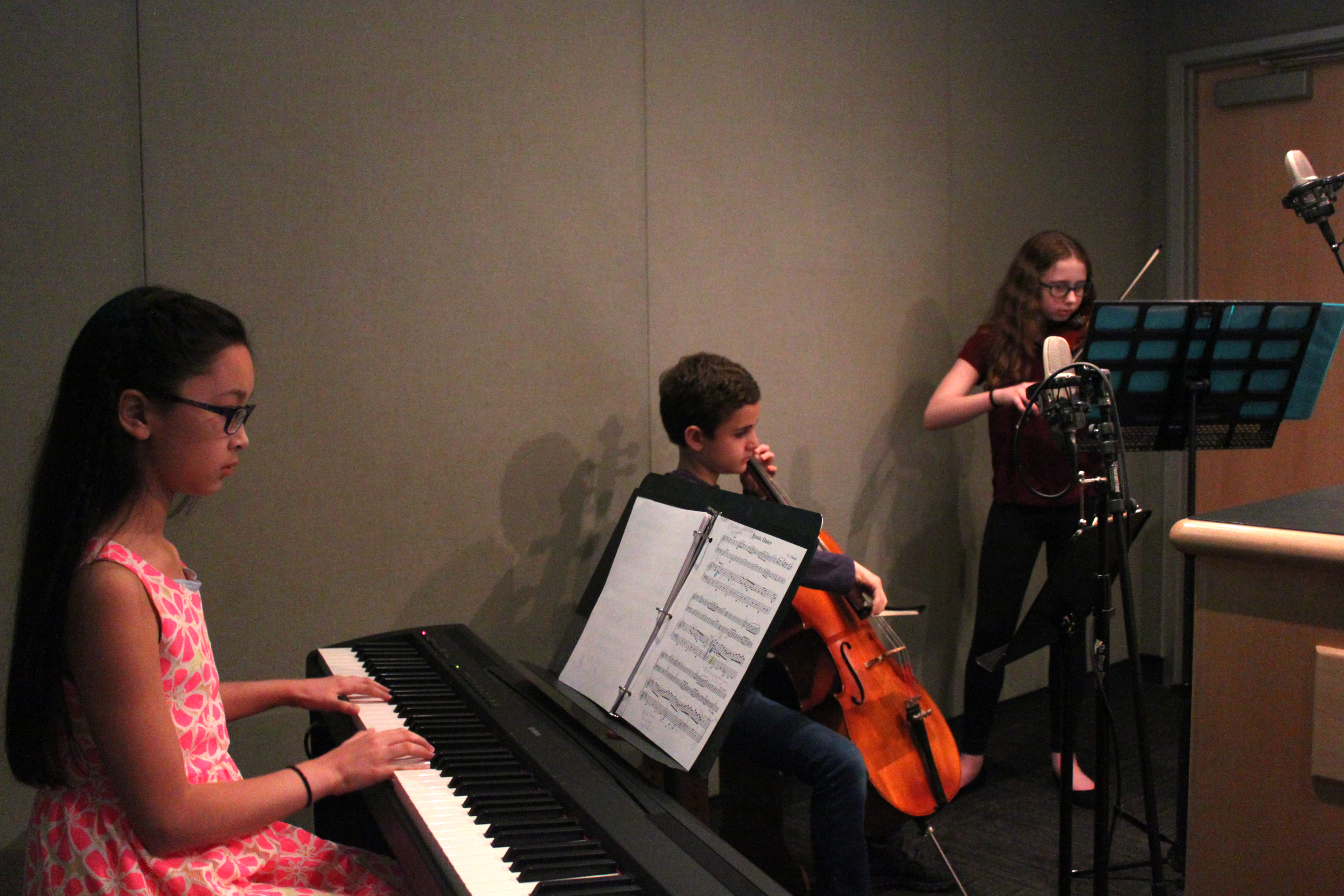 A child playing a piano keyboard, a child playing cello, and a child playing violin, all looking at sheet music on music stands in a radio studio