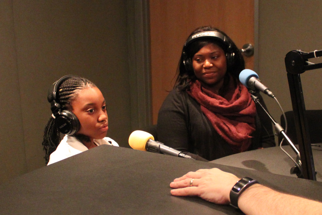 Two teens wearing headphones sitting behind microphones being interviewed in a radio studio