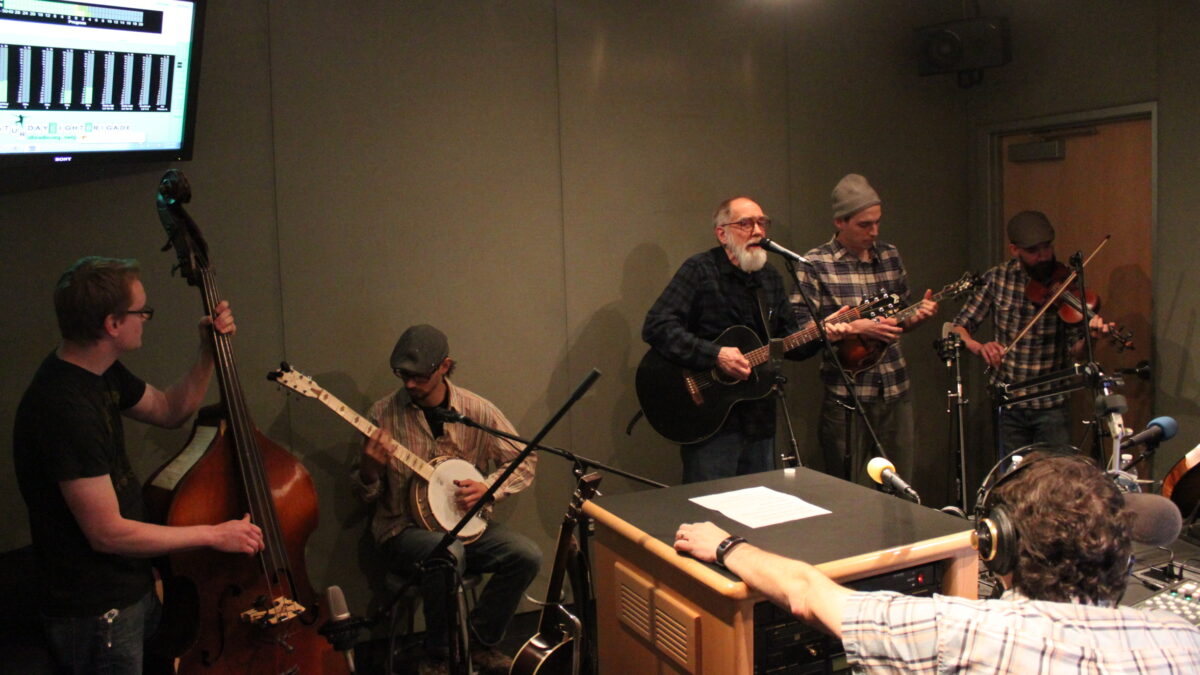 An adult playing upright bass, an adult playing banjo, an adult playing acoustic guitar and singing into a microphone, an adult playing mandolin, and an adult playing fiddle while an adult wearing headphones sitting behind a microphone listens in a radio studio