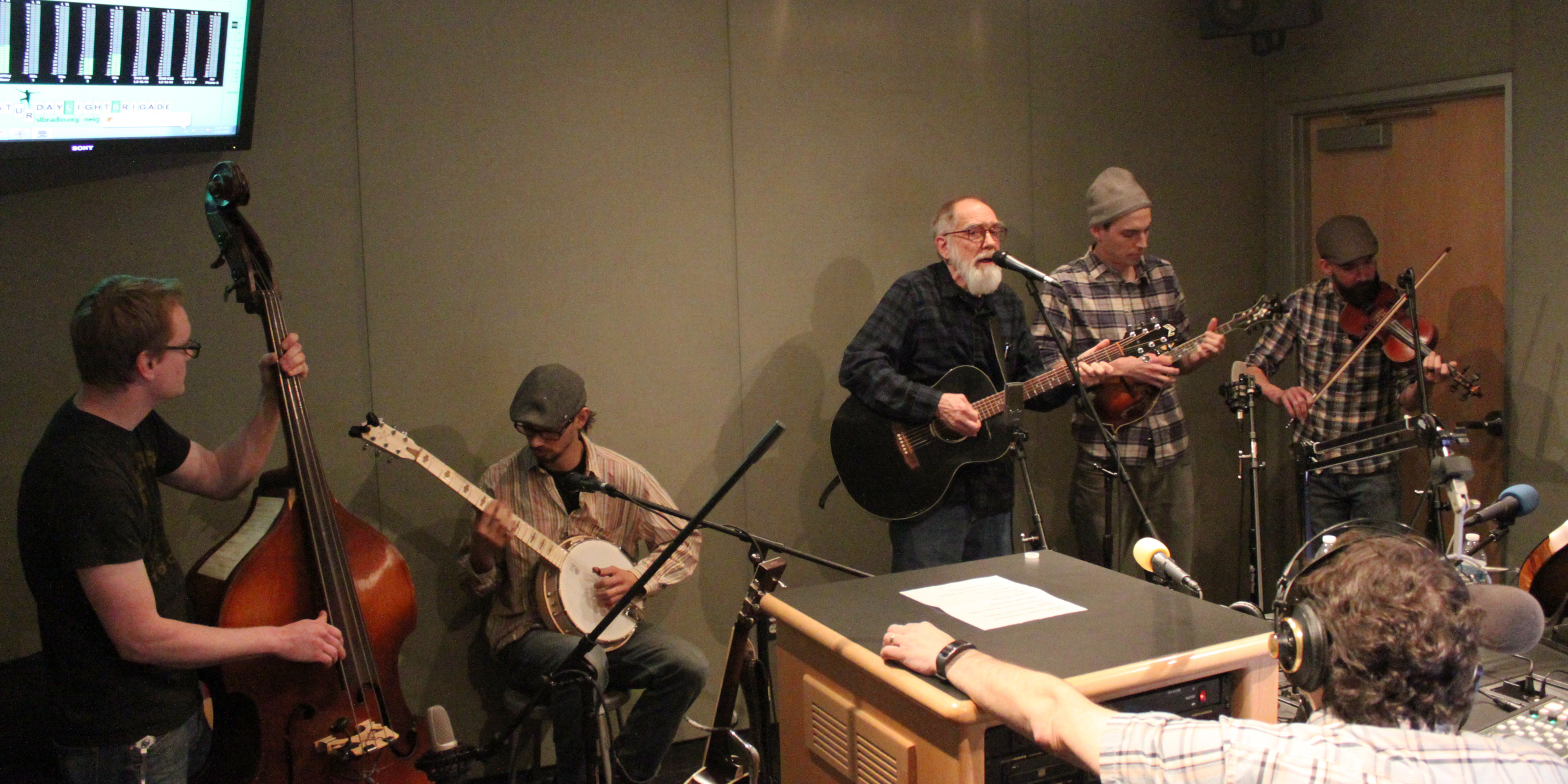 An adult playing upright bass, an adult playing banjo, an adult playing acoustic guitar and singing into a microphone, an adult playing mandolin, and an adult playing fiddle while an adult wearing headphones sitting behind a microphone listens in a radio studio