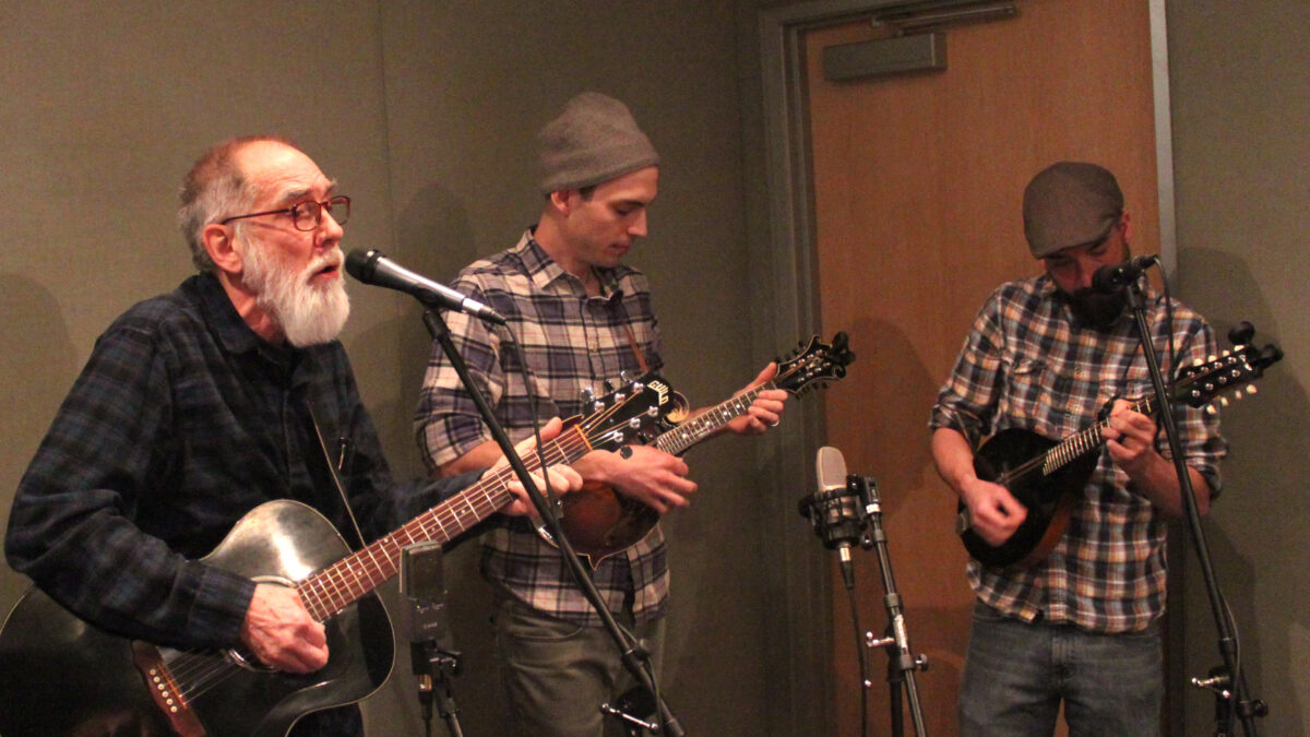An adult playing acoustic guitar and singing into a microphone and two adults playing mandolins in a radio studio