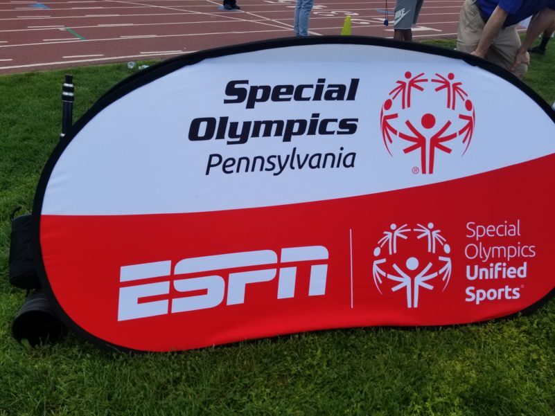 A bean shaped sign in a field of grass with the text "Special Olympics Pennsylvania" in black next to a red graphic of stick figures with multiple arms making a sphere all on a white background, and an ESPN logo with a white graphic of stick figures with multiple arms making a sphere and the text "Special Olympics Unified Sports" in white, all on a red background