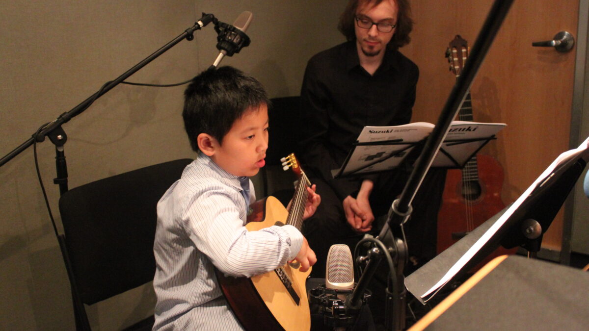 A youth looking at sheet music on a music stand and playing acoustic guitar and an adult in the background listening in a radio studio