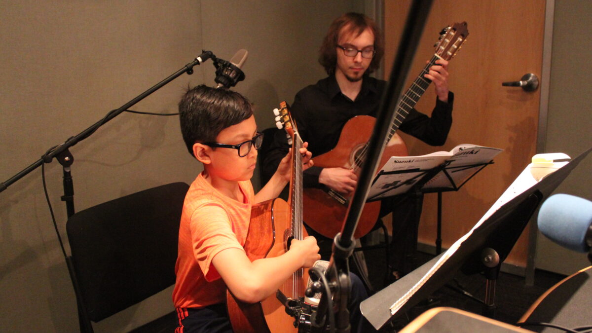 A youth looking at sheet music on a music stand and playing acoustic guitar and an adult in the background playing acoustic guitar and looking at sheet music on a music stand in a radio studio