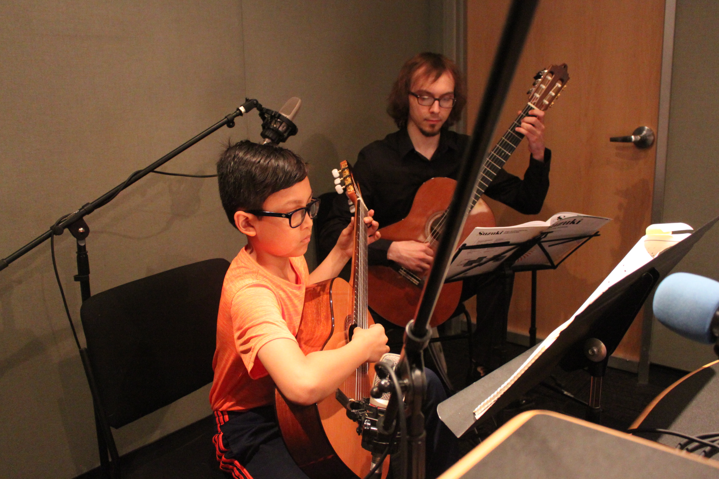 A youth looking at sheet music on a music stand and playing acoustic guitar and an adult in the background playing acoustic guitar and looking at sheet music on a music stand in a radio studio