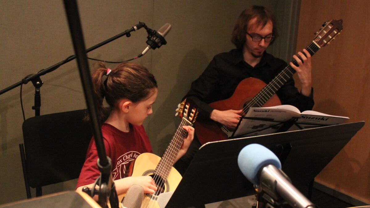 A youth looking down playing acoustic guitar and an adult in the background playing acoustic guitar and looking at sheet music on a music stand in a radio studio