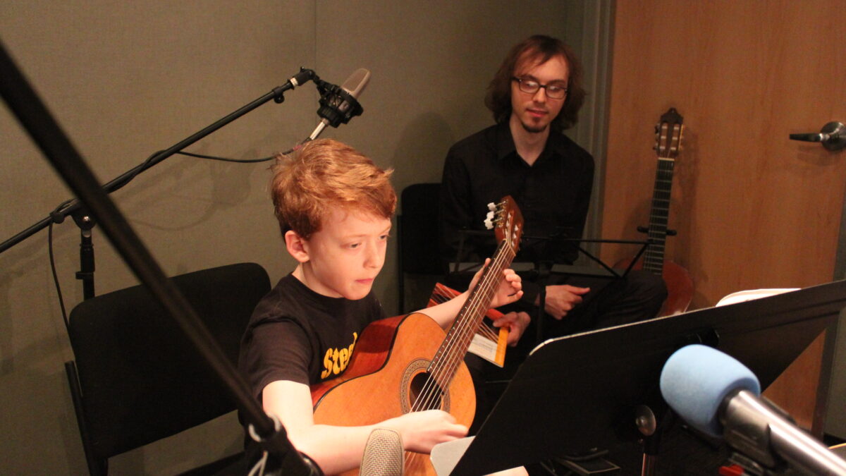 A youth playing acoustic guitar and looking at sheet music on a music stand while an adult sitting in the background listens in a radio studio