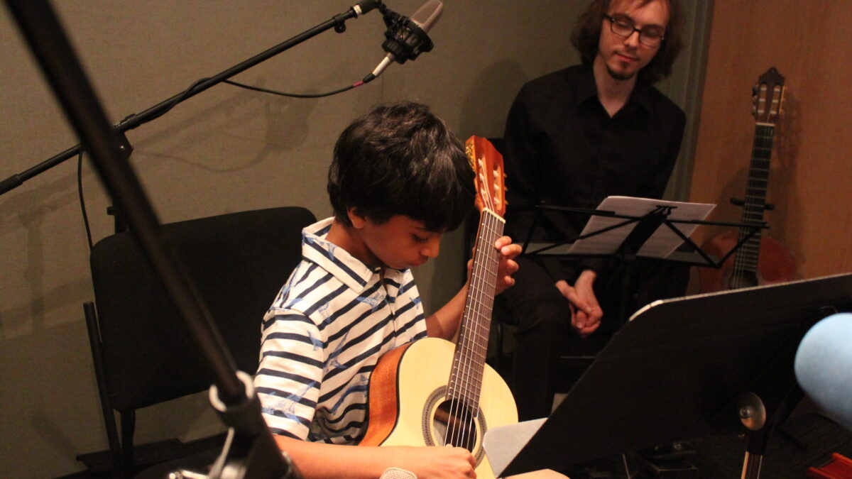 A youth looking down and playing an acoustic guitar as an adult watches in the background in a radio studio