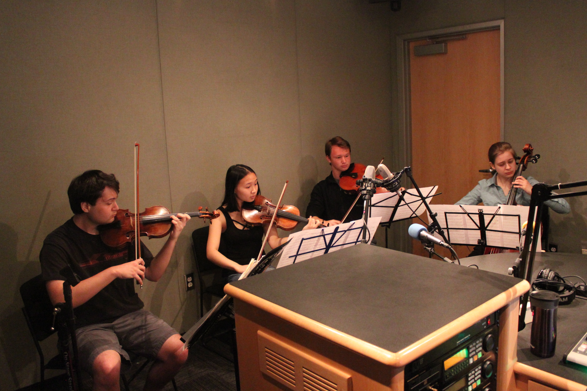 Two teens playing violin, one teen playing viola, and a teen playing cello, all looking at sheet music on music stands in a radio studio
