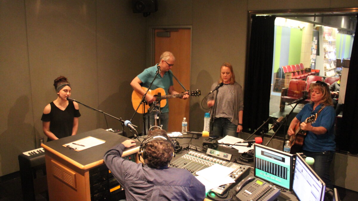 An adult playing a piano keyboard and singing into a microphone, an adult playing acoustic guitar, an adult singing into a microphone, and an adult playing acoustic guitar and singing into a microphone while an adult wearing headphones and sitting behind a microphone and a mixing board listens in a radio studio