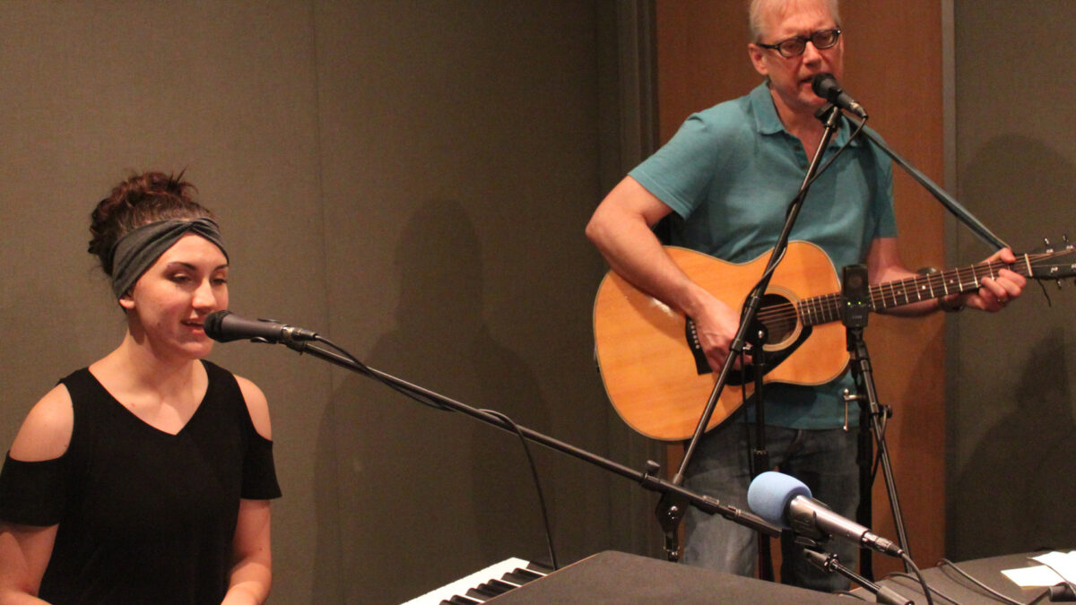 An adult playing a piano keyboard and singing into a microphone and an adult playing acoustic guitar and singing into a microphone in a radio studio