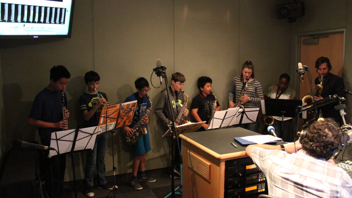 A youth playing clarinet, a youth playing trumpet, four youth playing alto saxophones, one youth playing tenor saxophone, and an adult playing baritone saxophone, all looking at sheet music on music stands, while an adult wearing headphones and sitting behind a microphone listens in a radio studio