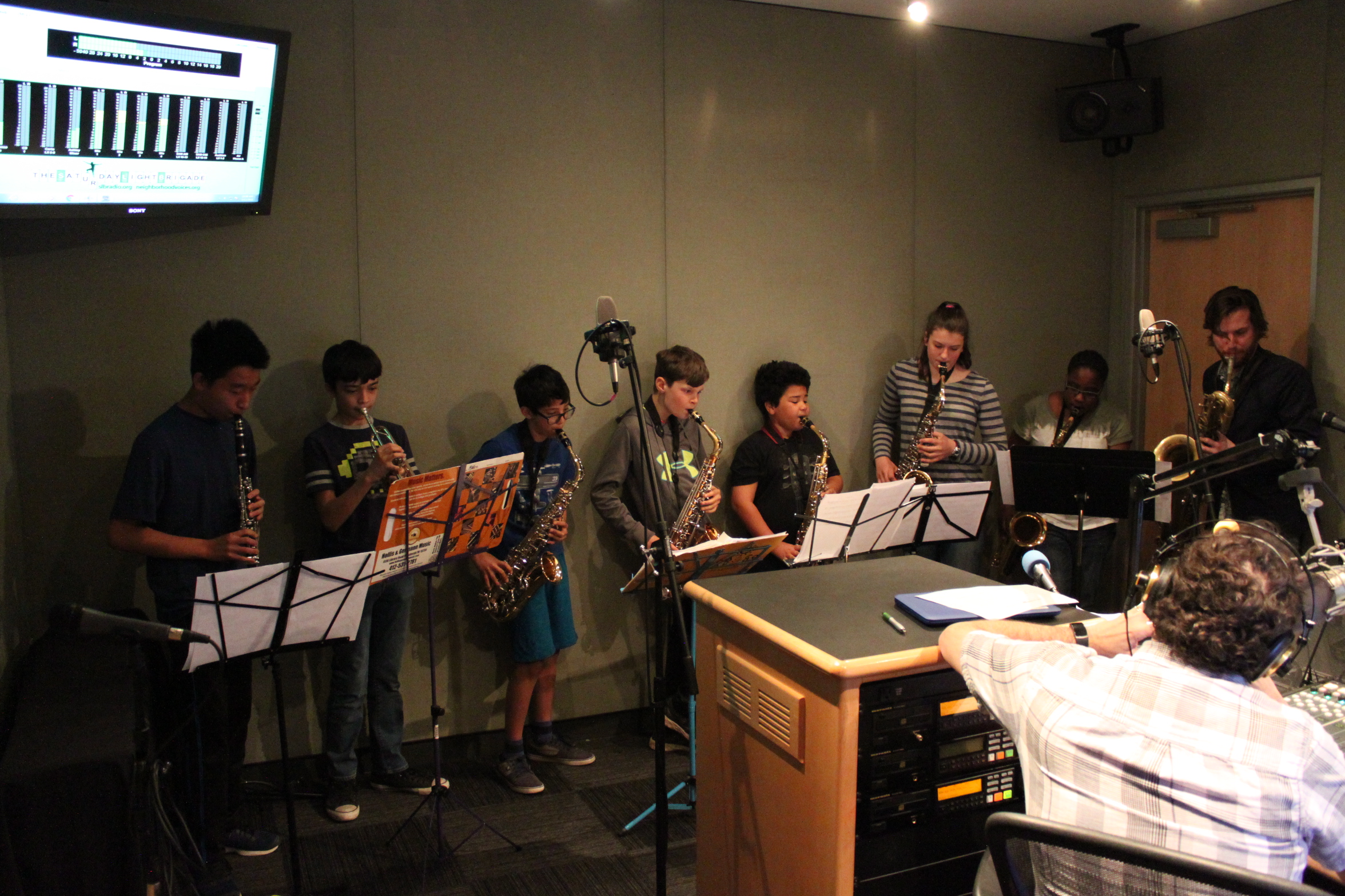 A youth playing clarinet, a youth playing trumpet, four youth playing alto saxophones, one youth playing tenor saxophone, and an adult playing baritone saxophone, all looking at sheet music on music stands, while an adult wearing headphones and sitting behind a microphone listens in a radio studio