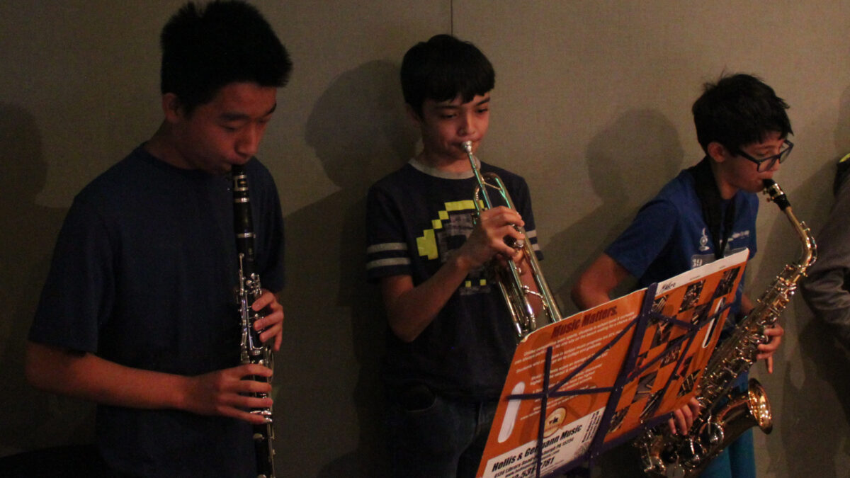 A youth playing clarinet, a youth playing trumpet, and a youth playing alto saxophone, all looking at sheet music on music stands in a radio studio