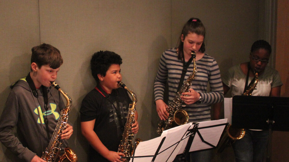 Three teens playing alto saxophone and one teen playing tenor saxophone, all looking at sheet music on music stands in a radio studio