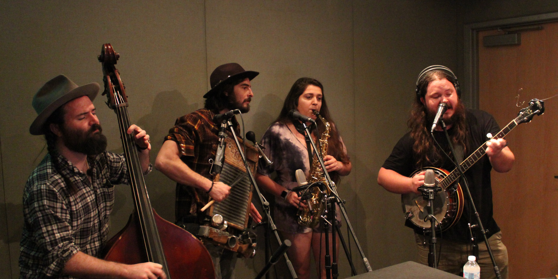 An adult playing upright bass, an adult playing a washboard, an adult playing alto saxophone, and an adult playing a banjo and singing into a microphone in a radio studio