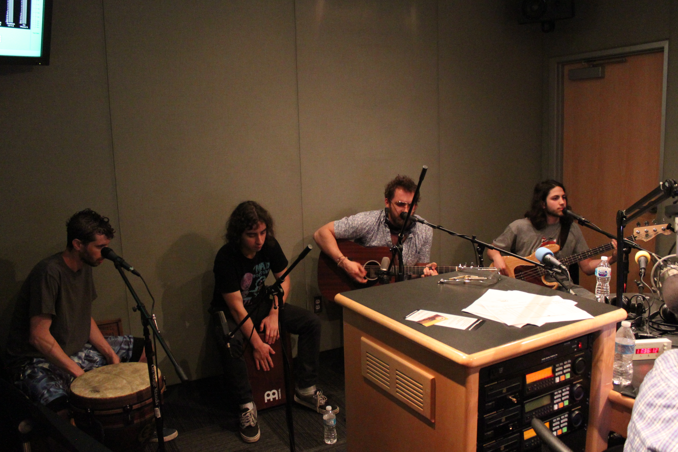 An adult playing a conga, an adult playing a djembe, an adult playing acoustic guitar and singing into a microphone, and an adult playing electric bass in a radio studio