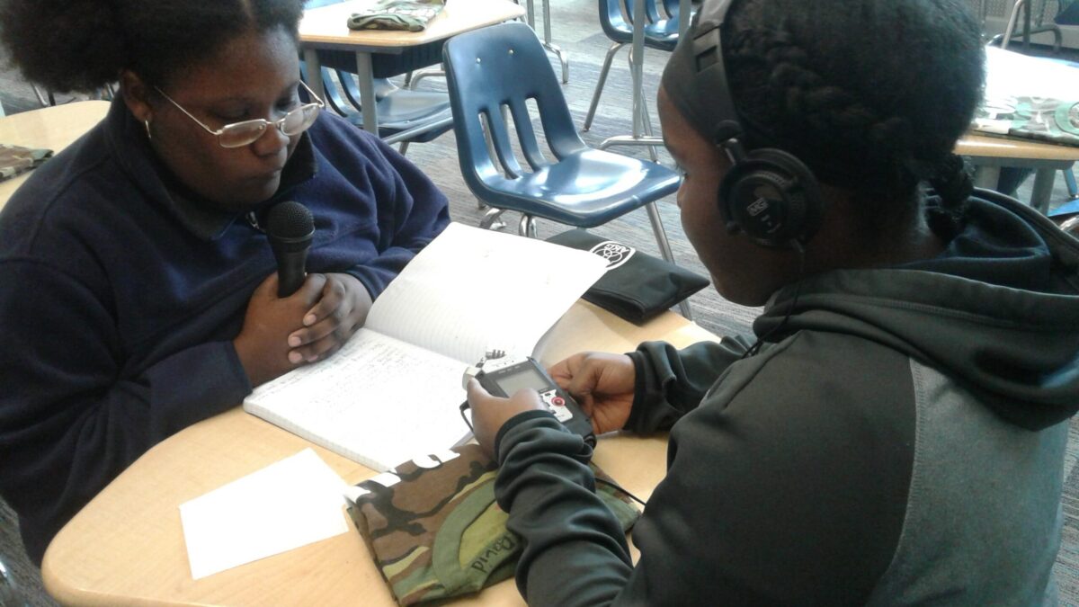 Teen wearing headphones holding a portable recorder interviewing another teen holding and speaking into a microphone
