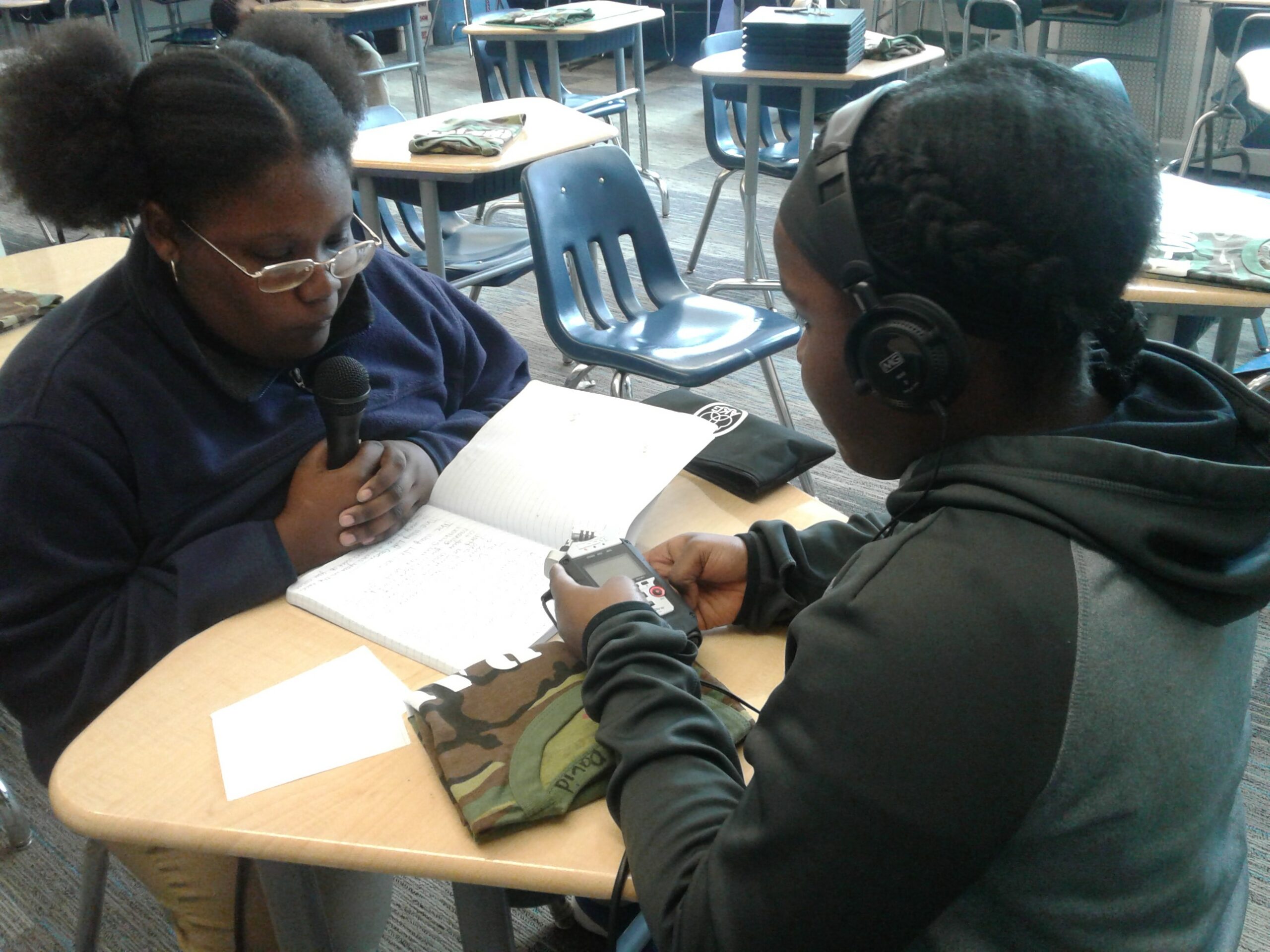 Teen wearing headphones holding a portable recorder interviewing another teen holding and speaking into a microphone