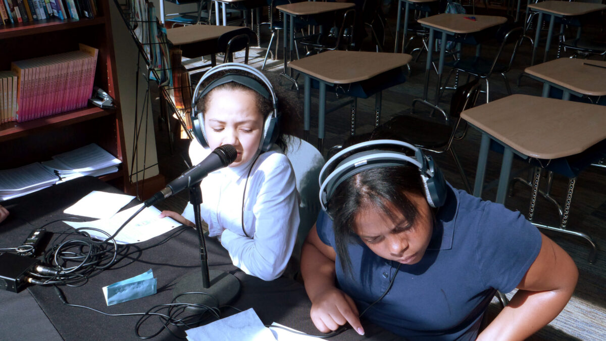 Two teens wearing headphones looking at pieces of paper on a black table, one speaking into a microphone