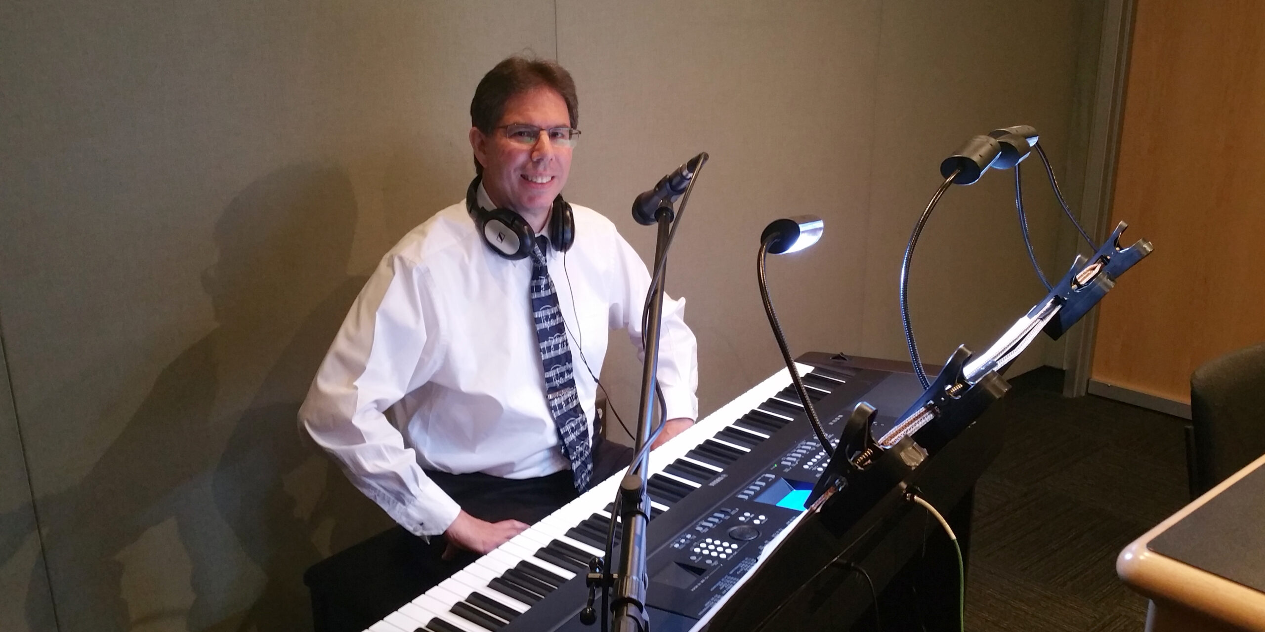 An adult wearing headphones smiling and sitting behind a piano keyboard in a radio studio