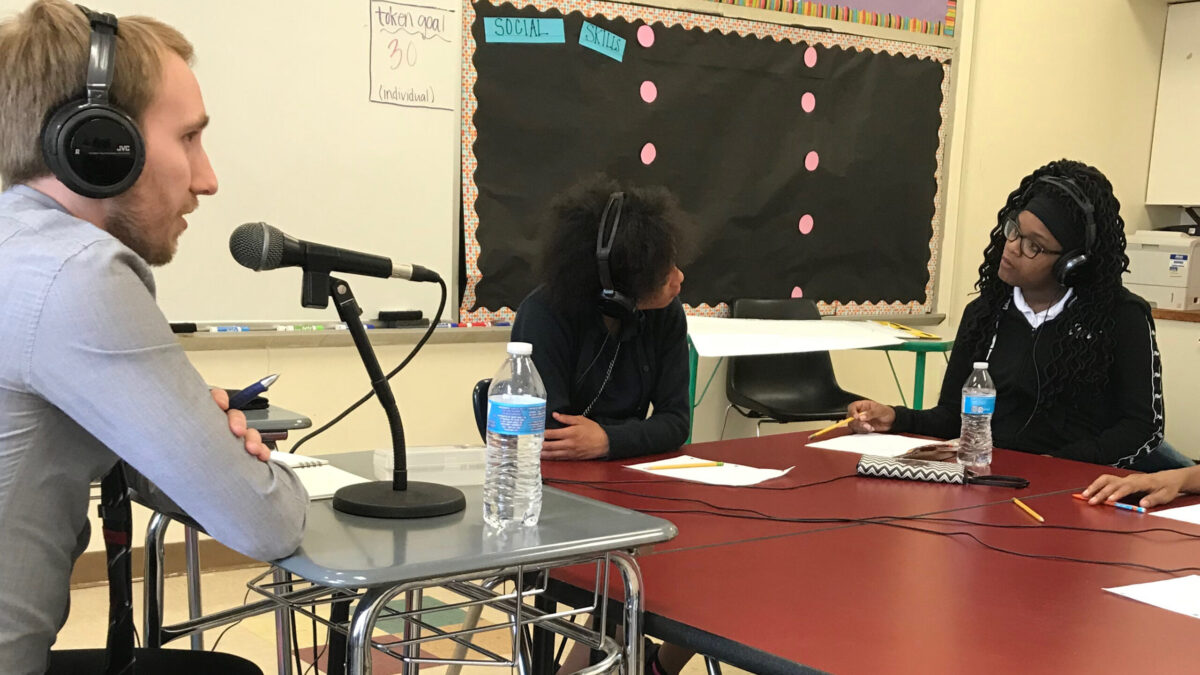 An adult wearing headphones speaking into a microphone to two teens wearing headphones looking at each other.
