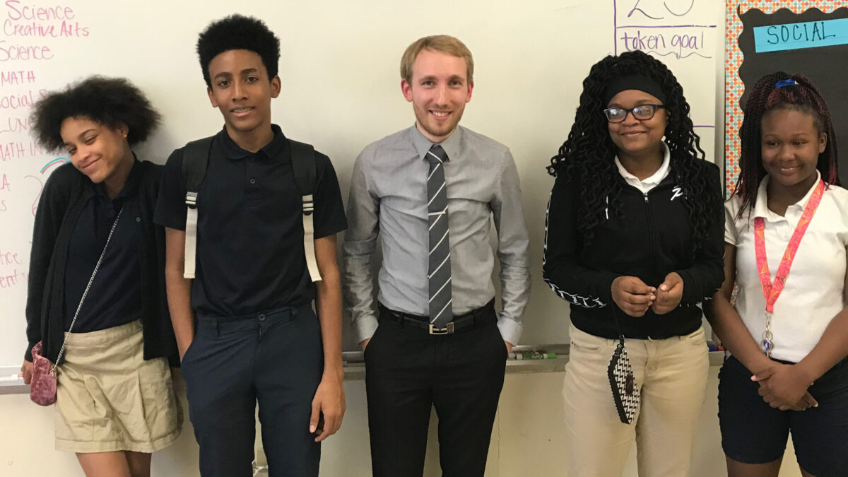 One adult and four youth smiling and posing in front of a white board in a classroom