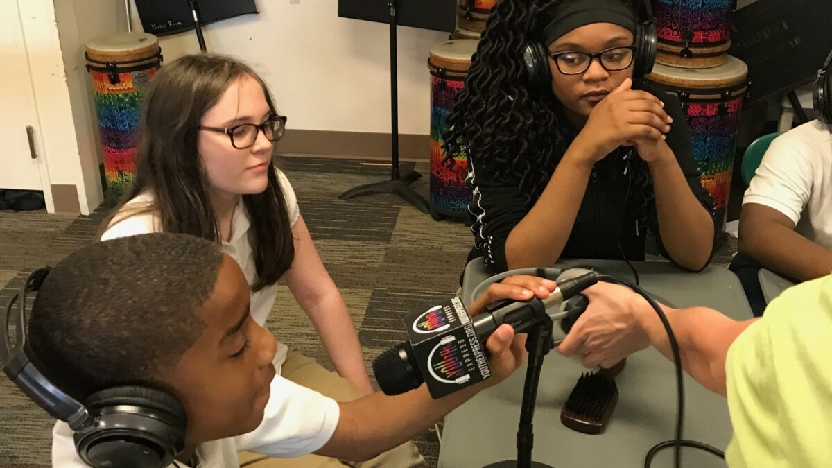 Three teens sitting around a gray table, two wearing headphones, one speaking into a microphone