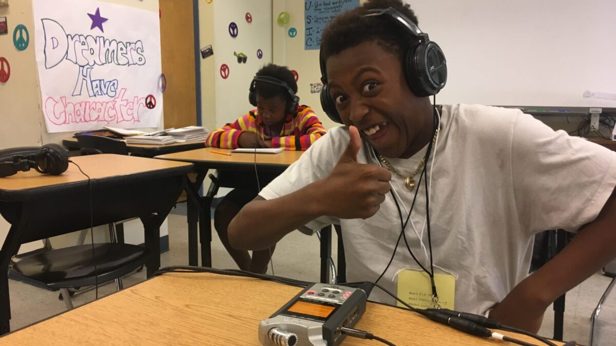 A teen wearing headphones smiling and giving a thumbs up while listening to a portable recorder on a wooden desk in a classroom, with another teen wearing headphones sitting at another desk in the background reading from a notepad