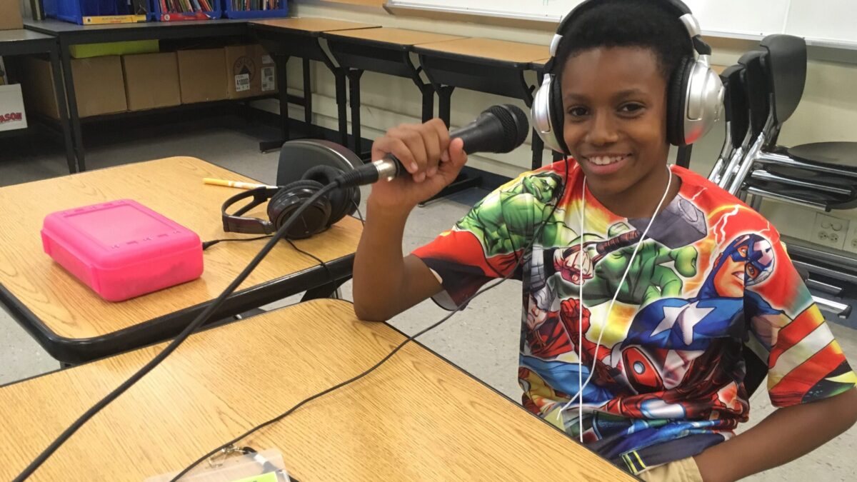 Teen wearing headphones smiling and holding a microphone sitting at a wooden desk in a classroom