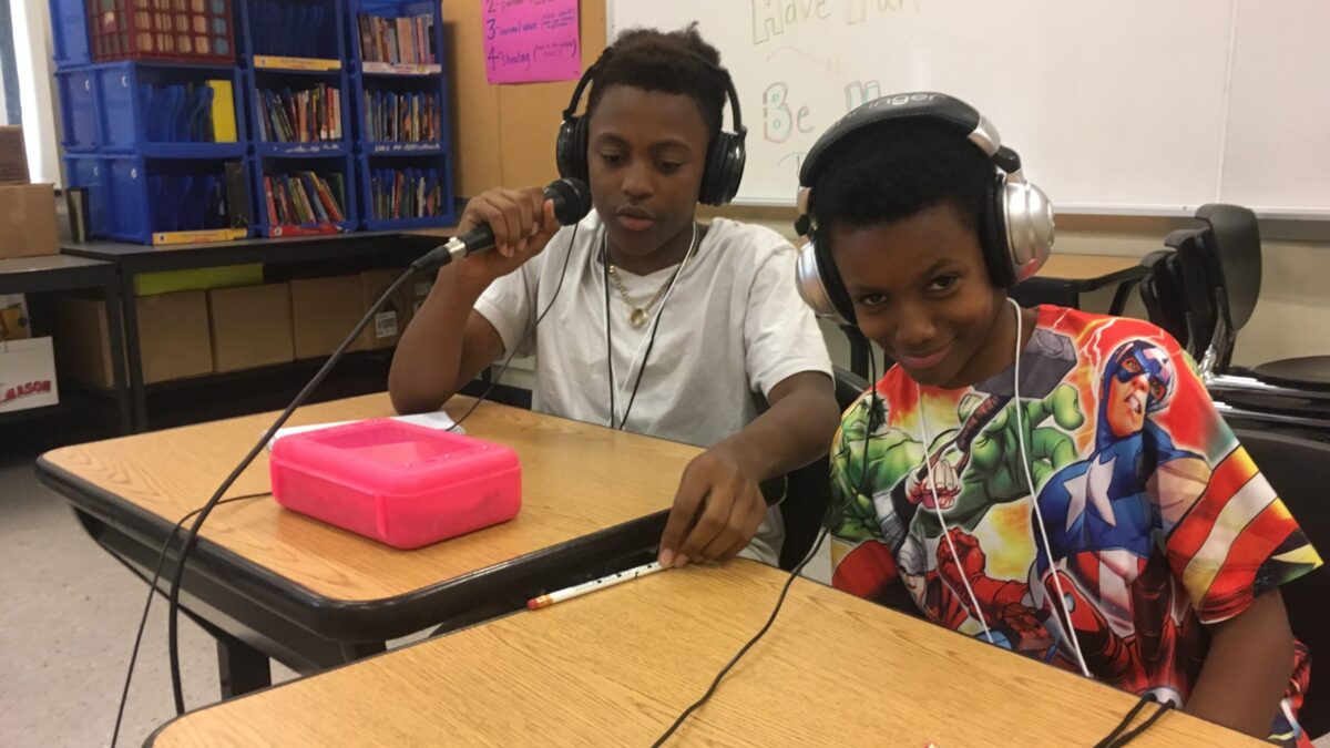 Two teens wearing headphones and sitting at wooden desks in a classroom, one holding a microphone