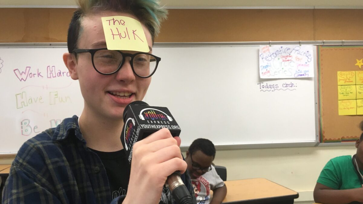 A teen with a sticky note reading "The Hulk" on their face speaking into a microphone standing in front of two teens sitting at wooden desks in front of a white board in a classroom