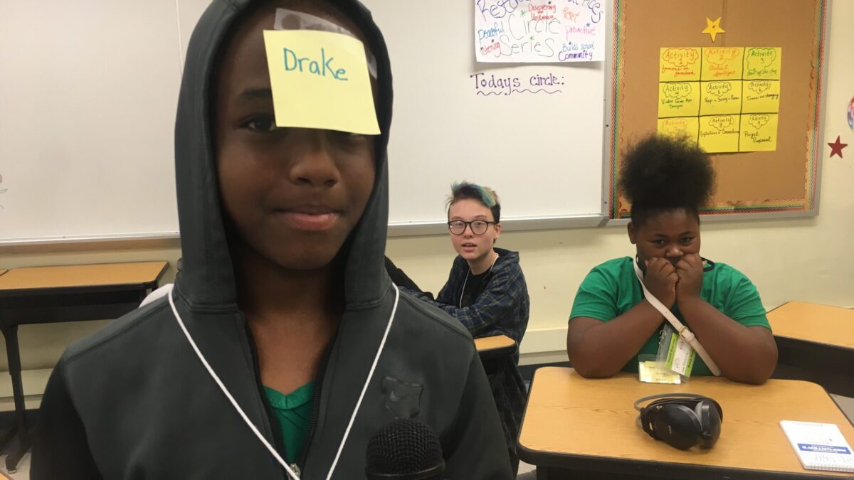 A teen with a sticky note reading "Drake" on their head holding a microphone as two teens look on behind them sitting at wooden desks in front of a white board in a classroom