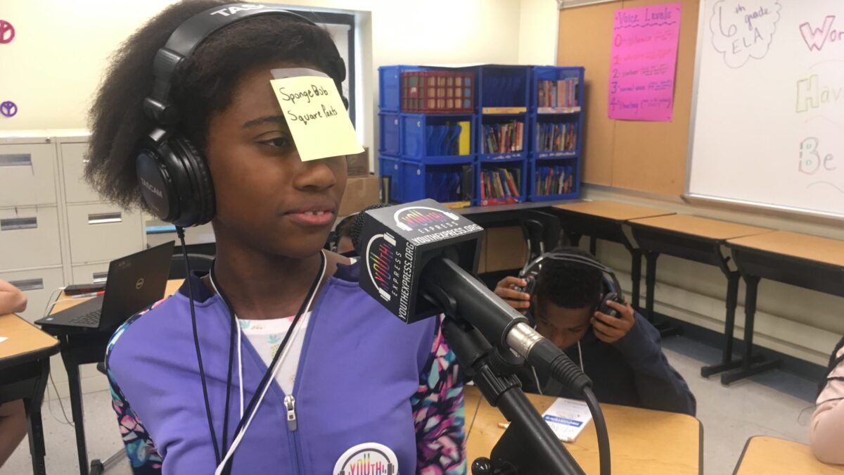 A teen with a sticky note on their face reading "Spongebob Squarepants" wearing headphones and speaking into a microphone in front of a teen wearing headphones sitting at a wooden desk in a classroom