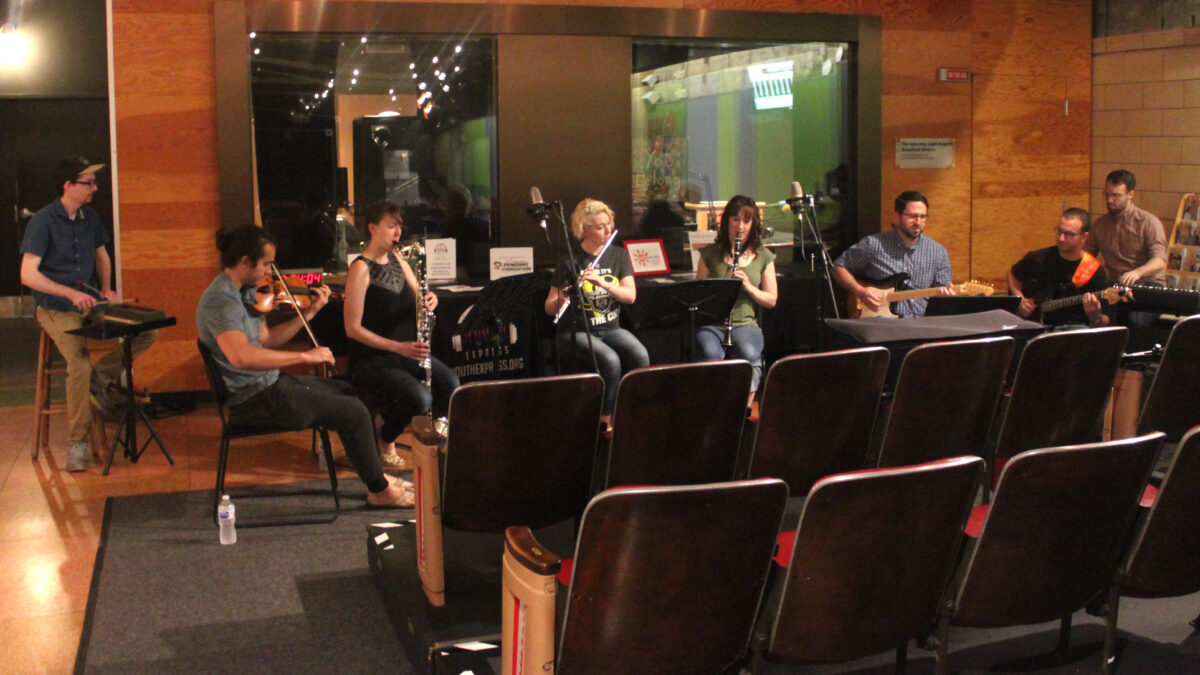 An adult playing wood block, an adult playing violin, an adult playing bass clarinet, an adult playing flute, an adult playing clarinet, two adults playing electric guitar, and an adult playing a piano keyboard