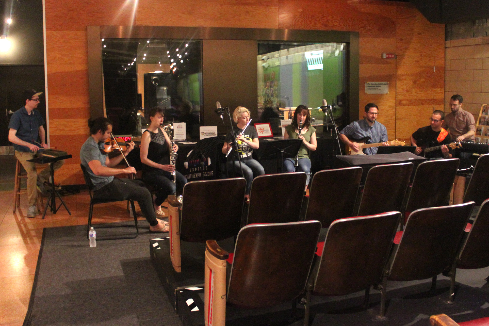 An adult playing wood block, an adult playing violin, an adult playing bass clarinet, an adult playing flute, an adult playing clarinet, two adults playing electric guitar, and an adult playing a piano keyboard