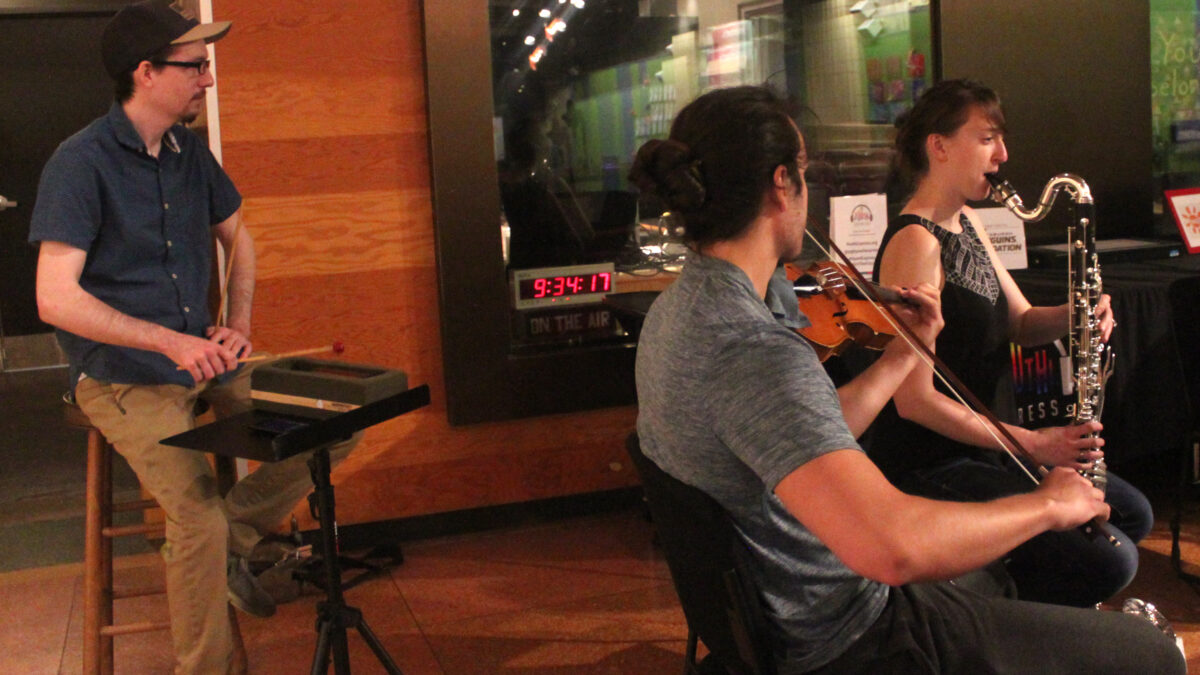 An adult playing a wood block, an adult playing violin, and an adult playing bass clarinet
