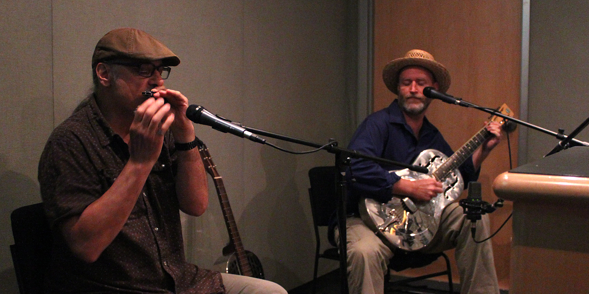 An adult playing harmonica into a microphone and an adult playing acoustic guitar sitting behind a microphone in a radio studio