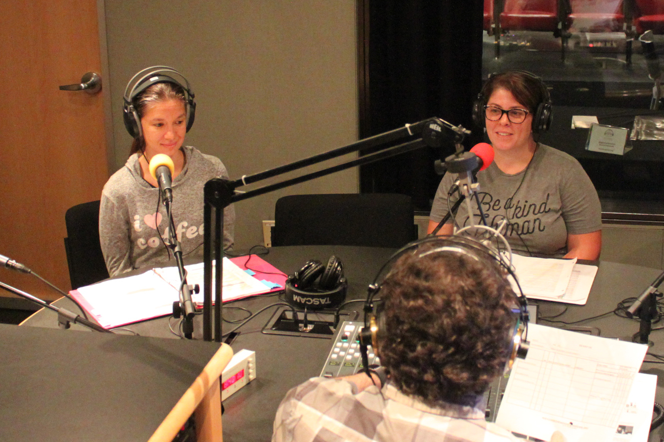 A teen and an adult wearing headphones and speaking into microphones being interviewed by an adult wearing headphones and sitting behind a microphone in a radio studio