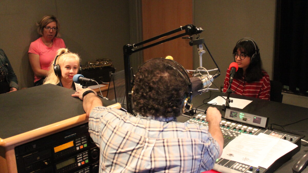 Two youth wearing headphones sitting behind microphones being interviewed by an adult wearing headphones and sitting behind a microphone and a mixing board in a radio studio as another adult listens in the background