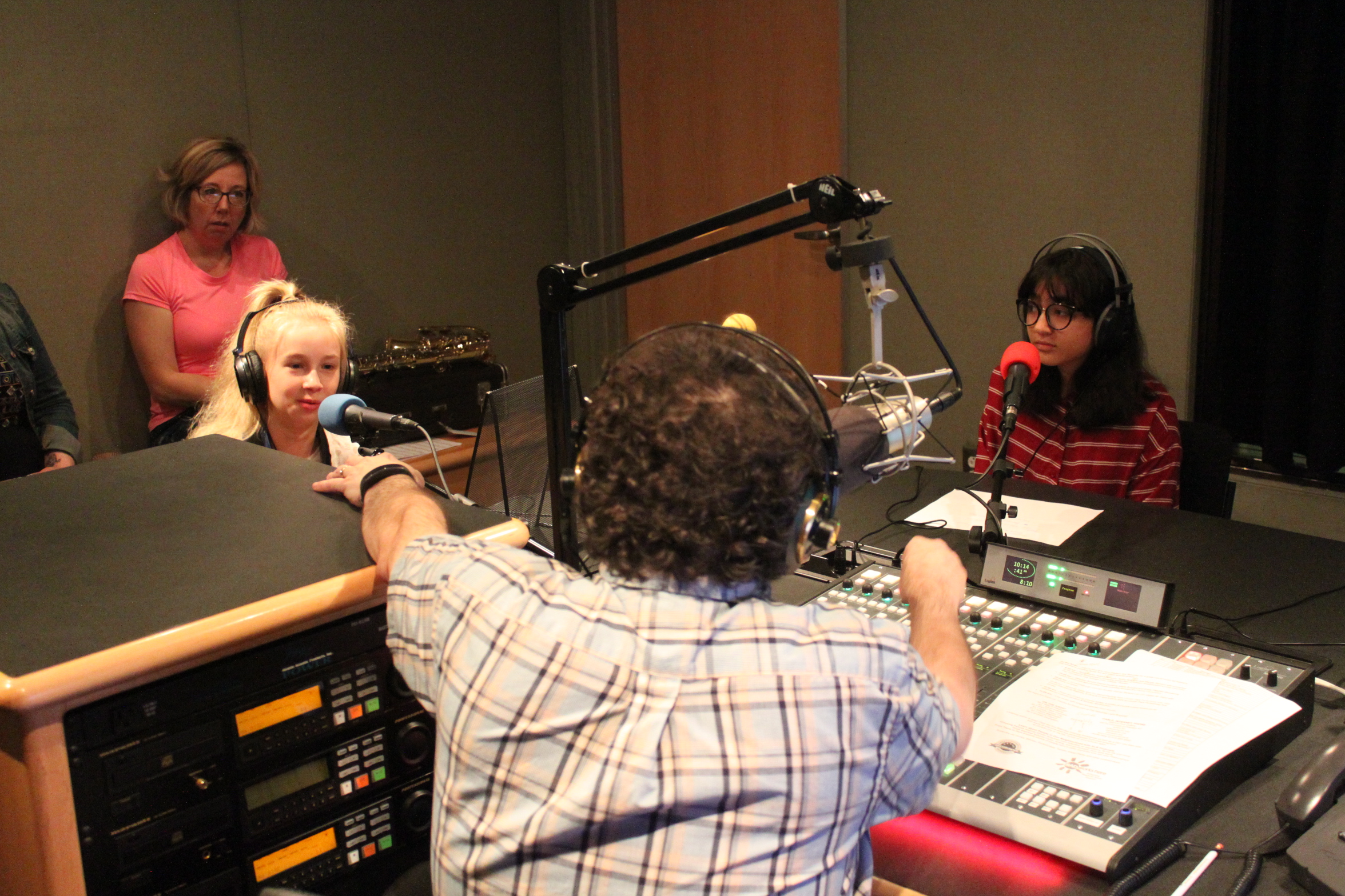 Two youth wearing headphones sitting behind microphones being interviewed by an adult wearing headphones and sitting behind a microphone and a mixing board in a radio studio as another adult listens in the background