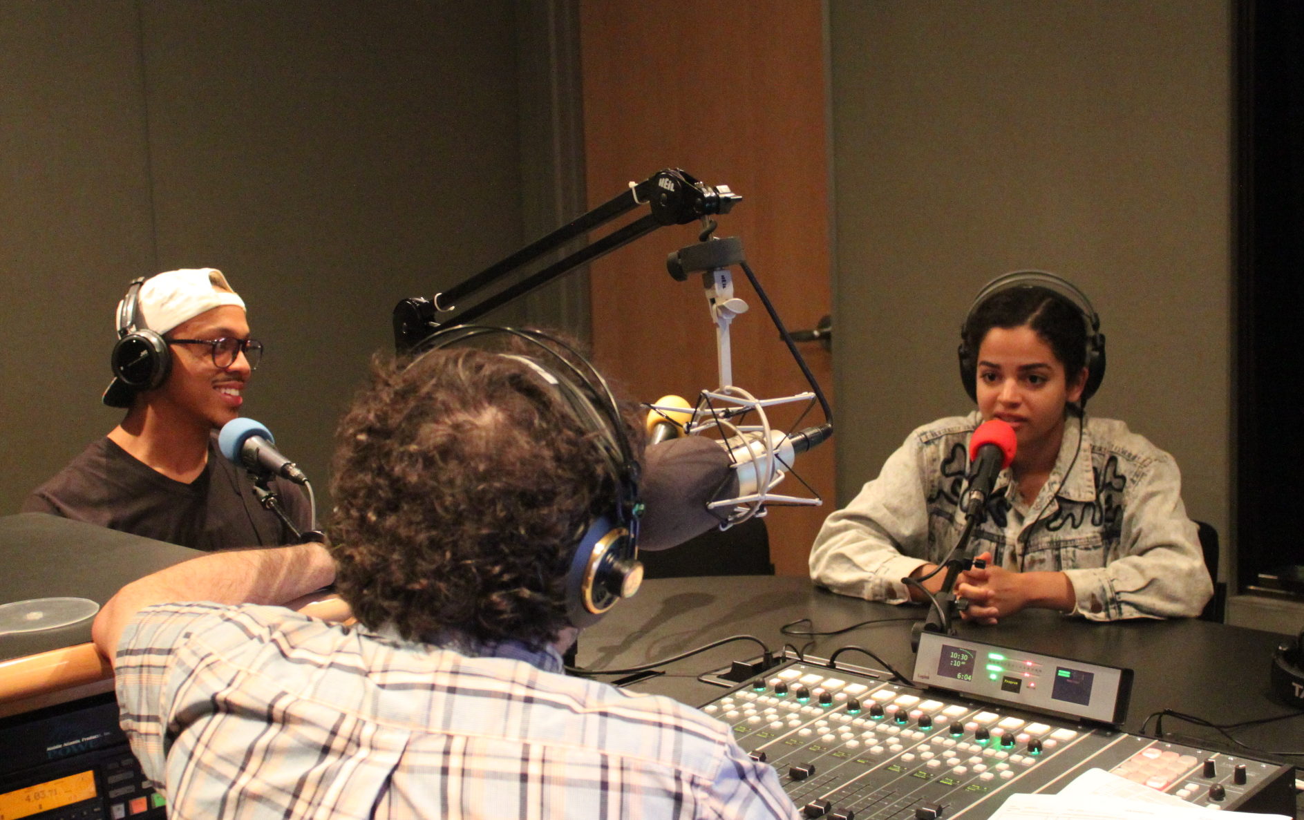 Two teens wearing headphones and speaking into microphones being interviewed by an adult wearing headphones and sitting behind a microphone and a mixing board in a radio studio