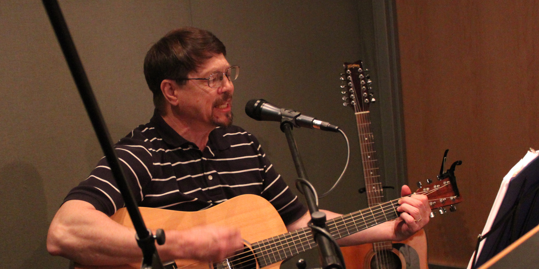 An adult playing acoustic guitar and singing into a microphone in a radio studio