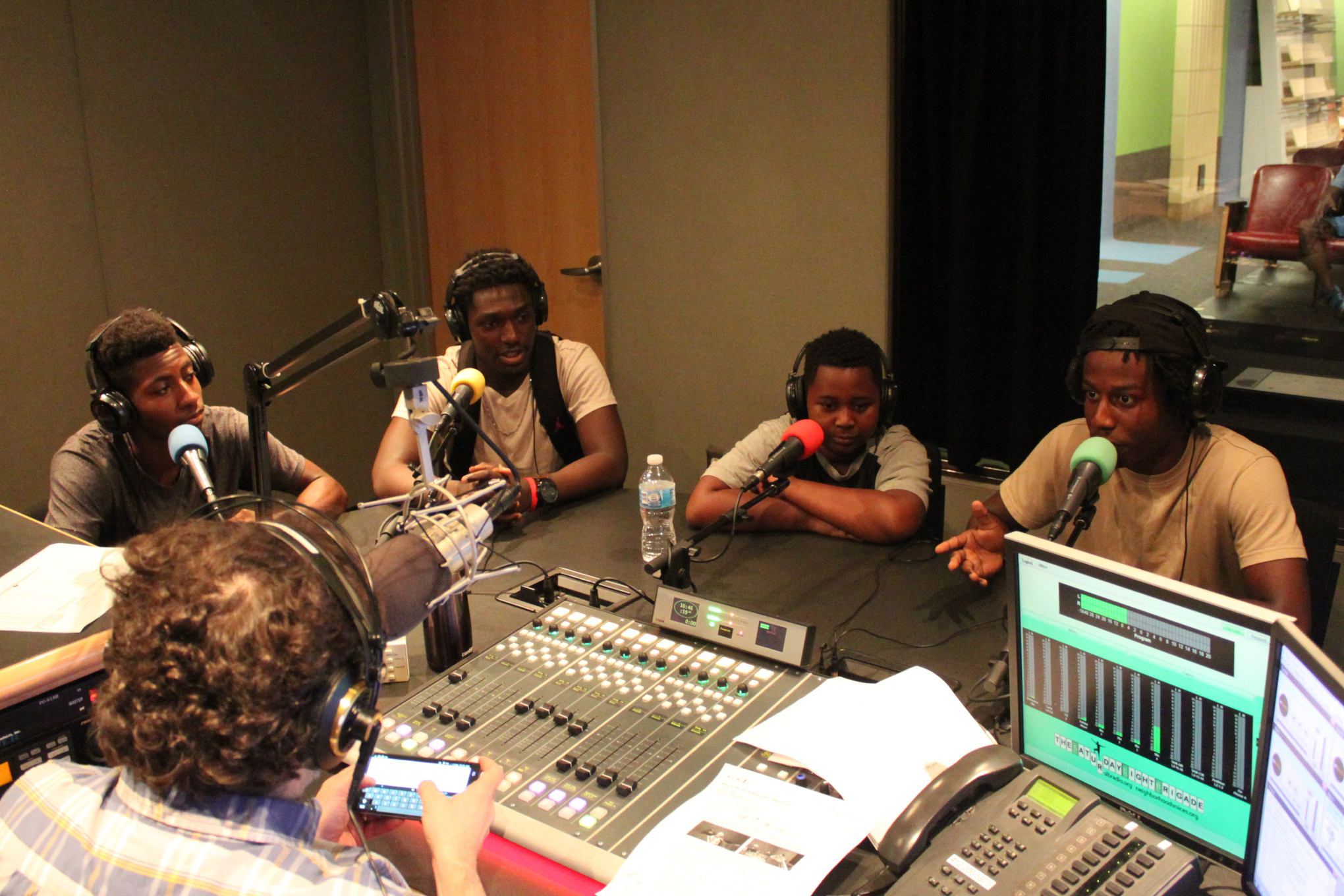 Four teens wearing headphones speaking into microphones being interviewed by an adult wearing headphones sitting behind a microphone and a mixing board in a radio studio
