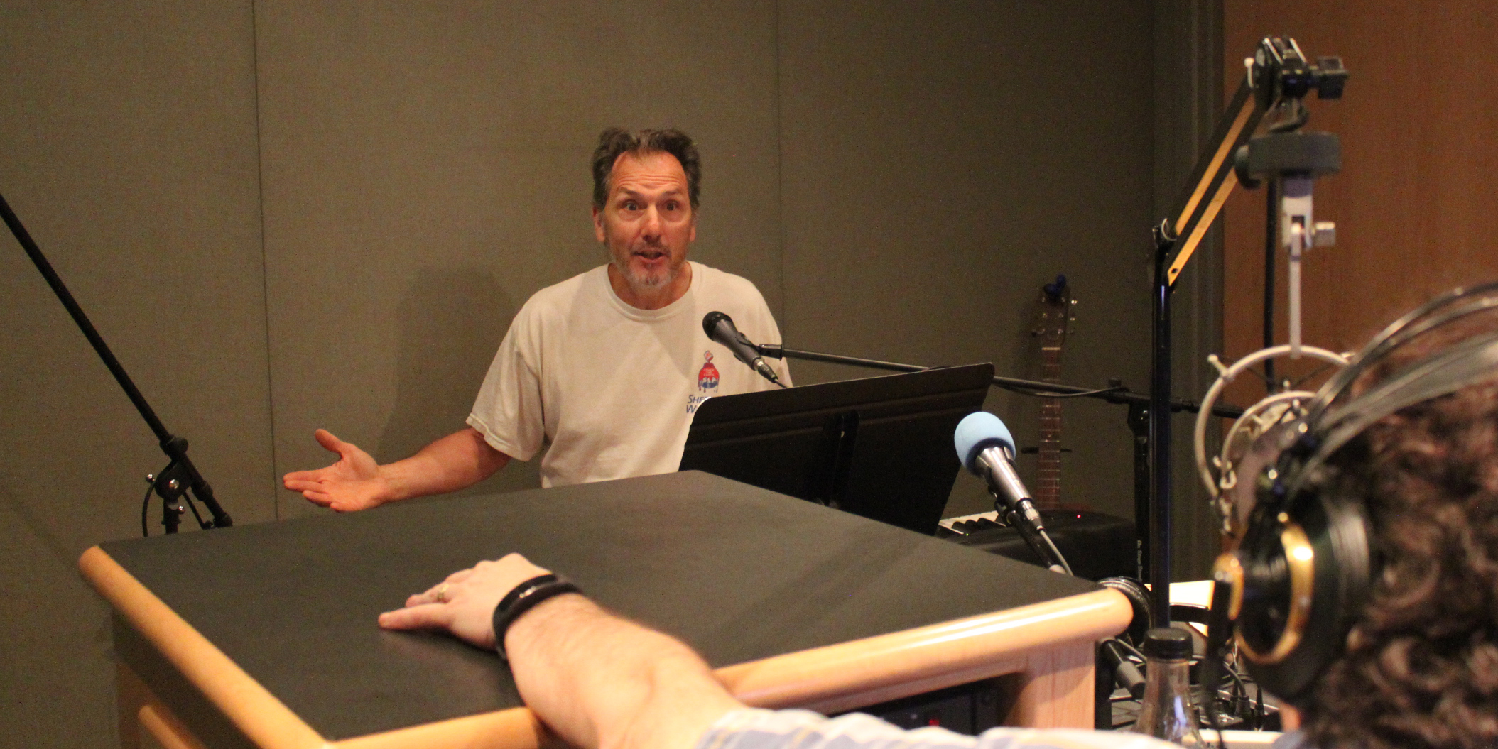 An adult sitting behind a piano keyboard speaking into a microphone and gesturing with their hands being interviewed by an adult wearing headphones in a radio studio