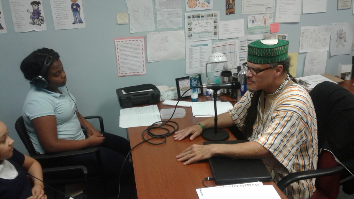 Two teens wearing headphones interviewing an adult sitting behind a desk speaking into a microphone