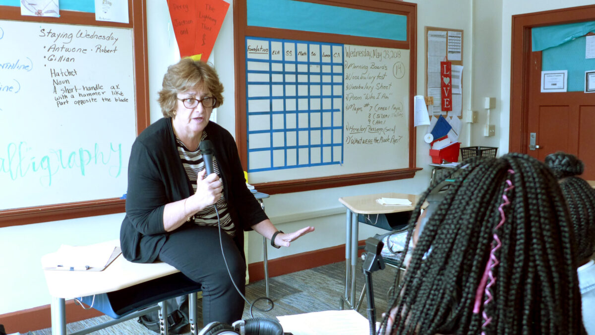 An adult sitting on a desk speaking into a microphone to two individuals sitting at classroom desks