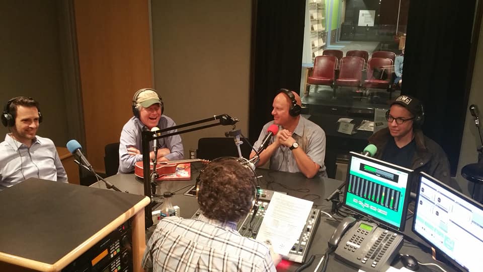 Four adults wearing headphones speaking into microphones being interviewed by an adult wearing headphones siting behind a microphone and a mixing board in a radio studio