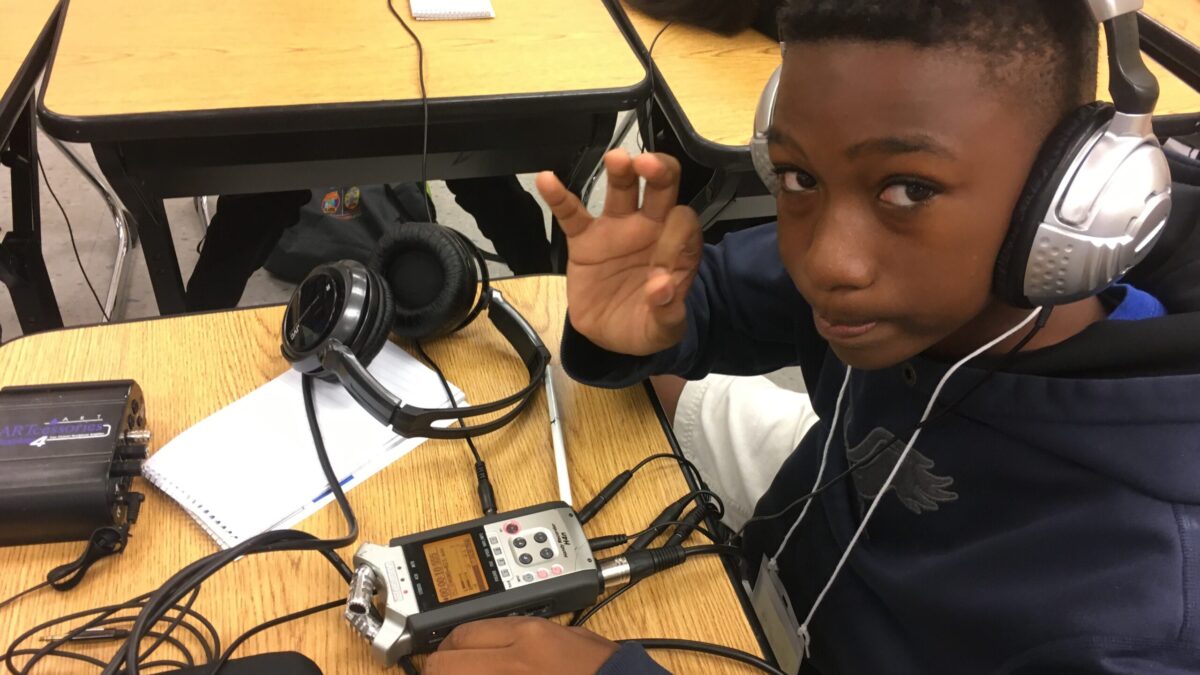 A youth wearing headphones listening to a portable recorder on a wooden table and making a circle with their fingers